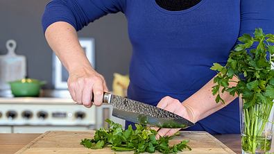 Eine Person hackt mit einem großen Damastmesser frische Petersilie auf einem Holzbrett in einer Küche. - Foto: LECKER @ Bauer Media Group / Emily Beckmann