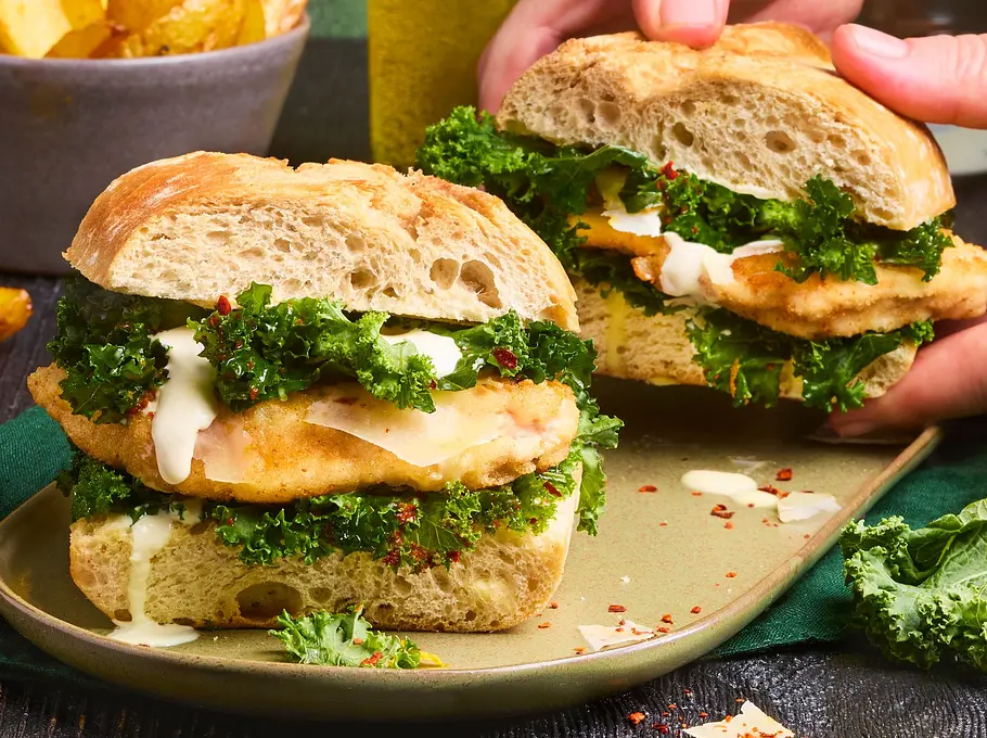Krosses Ciabatta-Sandwich gefüllt mit gebratenem Hähnchenfilet, frischem Grünkohl und cremiger Sauce, serviert auf einem grünen Teller mit goldenen Pommes in einer Schale im Hintergrund.