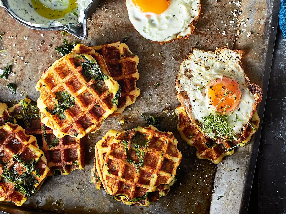 Goldbraune Kartoffel-Quark-Waffeln mit Spinat und Spiegelei liegen auf einem Blech, daneben ein Töpfchen mit Kräuterquark.