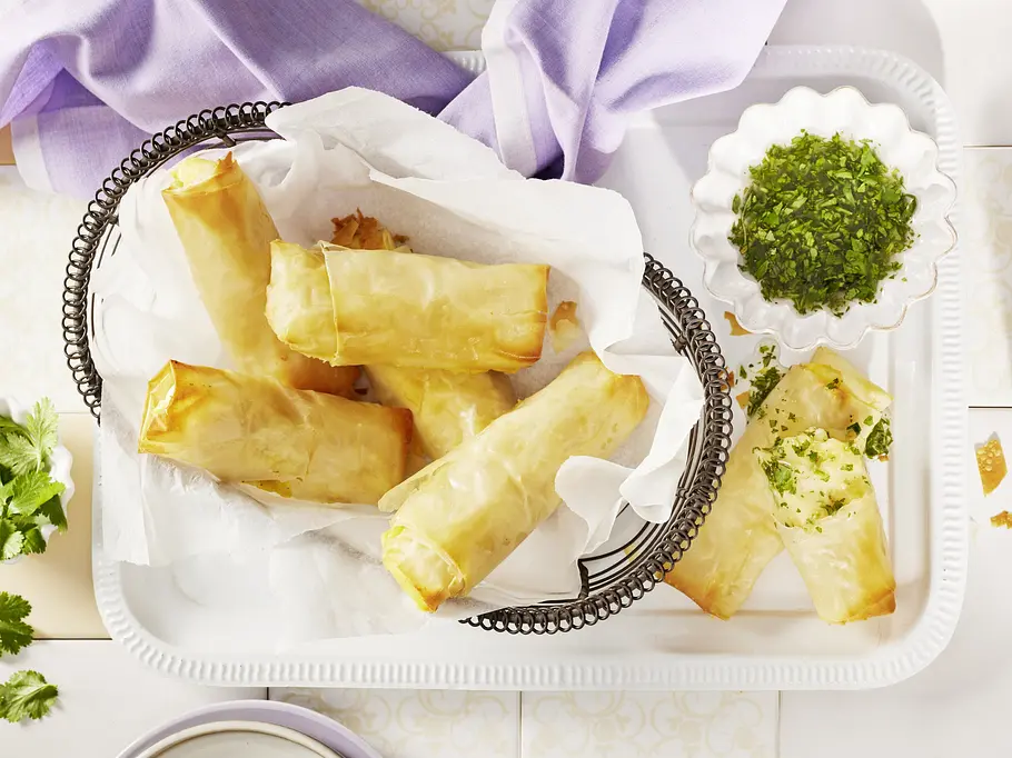 Kartoffelpüree-Röllchen mit Petersilienöl Goldbraune Teigrollen liegen in einem mit Papier ausgelegten Drahtkorb. Daneben steht eine Schale mit gehackten Kräutern.