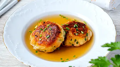 In einem weißen Suppenteller liegen zwei Kaspressknödel als Einlage in einer Brühe, garniert mit Schnittlauch - Foto: ShowHeroes für LECKER @ Bauer Media Group