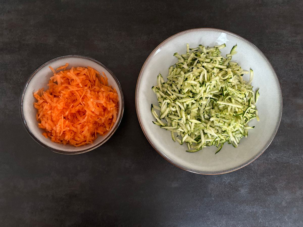 Zwei Schüsseln mit geraspelter Zucchini und Karotte auf dunklem Untergrund