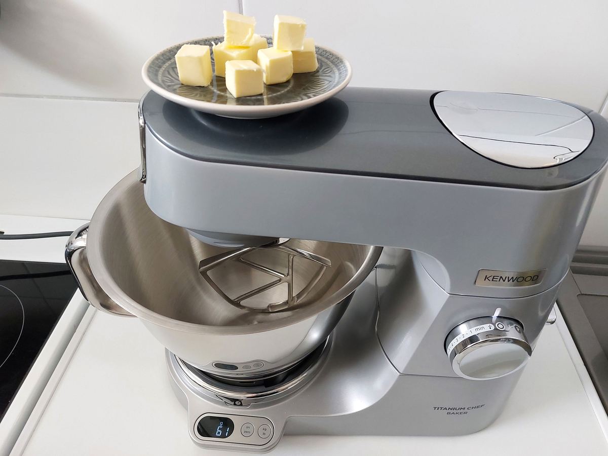 Eine graue Kenwood-Küchenmaschine mit Edelstahlschüssel steht auf der Arbeitsfläche, obenauf liegt ein Teller mit Butterwürfeln.