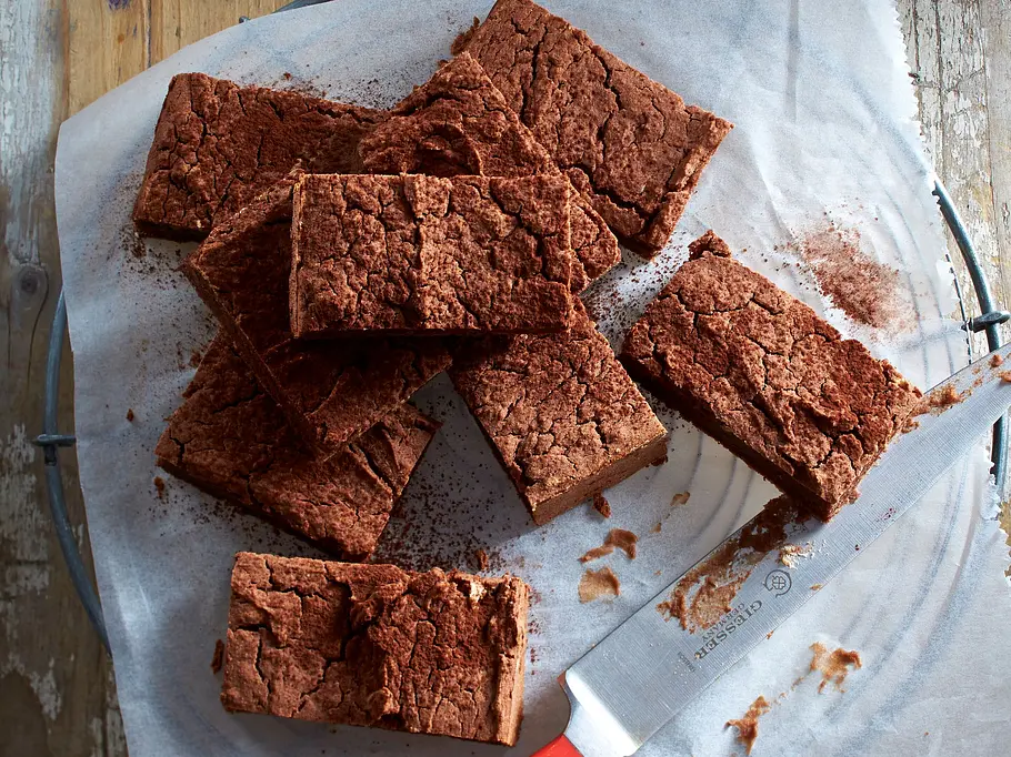 Kidneybohnen-Brownies mit Pekannüssen Rezept Mehrere dunkle, saftige Brownie-Stücke mit crackliger Oberfläche liegen auf einem Stück Backpapier, daneben ein Messer mit rotem Griff, serviert auf einem rustikalen Holztisch.