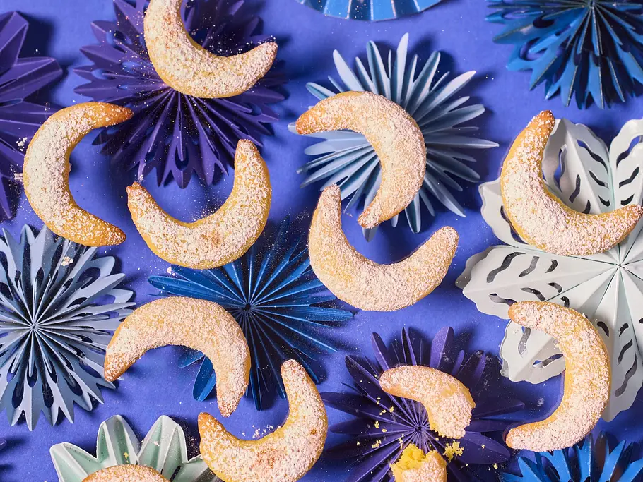 Halbmondförmige Kipferl „Golden Milk“, bestäubt mit gelbem Puder, auf blauen und lilafarbenen Papierstern-Unterlagen auf lilafarbener Oberfläche angerichtet.