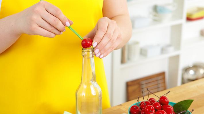 Eine Kirsche liegt zum Entkernen auf der Öffnung einer Glasflasche. Mit einem grünen Essstäbchen wird oben in die Kirsche gestochen, um den Kern unten herauszudrücken. - Foto: House of Food / Bauer Food Experts KG