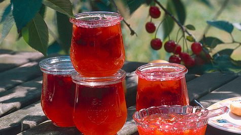 Mehrere Gläser mit frisch eingekochter Kirschmarmelade auf einem Holztisch im Garten, im Hintergrund hängen reife Kirschen am Baum. - Foto: House of Food / Bauer Food Experts KG