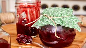 Ein Glas Kirschmarmelade mit grünem Stoffdeckel steht neben einem geöffneten Glas und einem Löffel auf einem Holzbrett. - Foto: House of Food / Bauer Food Experts KG