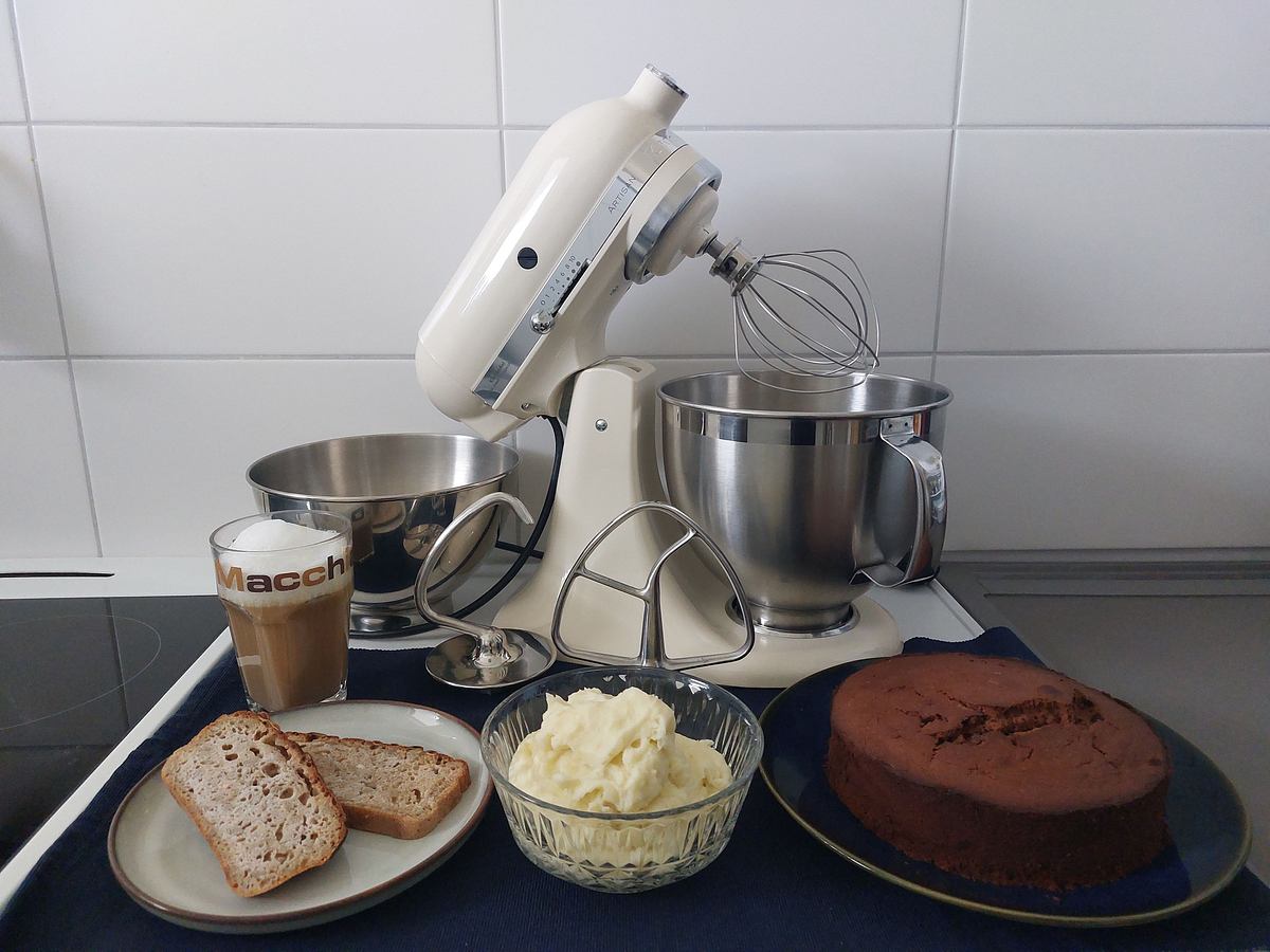 Eine cremefarbene KitchenAid-Küchenmaschine steht mit Rührschüsseln und Rührwerkzeugen auf der Arbeitsfläche, davor Brot, Kartoffelpüree, Kuchen und ein Glas Latte Macchiato mit Schaumkrone.