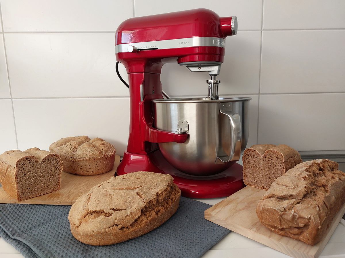 Eine rote KitchenAid 6,6 l Küchenmaschine steht auf einer Arbeitsfläche, umgeben von mehreren frisch gebackenen Broten auf Holzbrettchen.