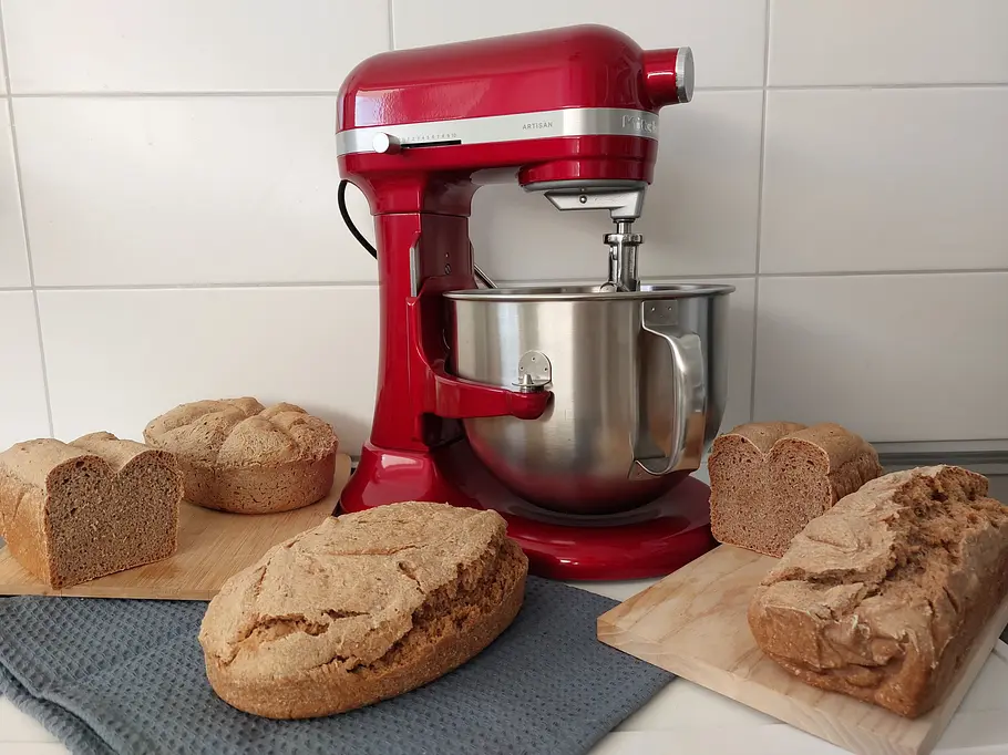 KitchenAid Artisan 6,6 l mit vier Broten Eine rote KitchenAid 6,6 l Küchenmaschine steht auf einer Arbeitsfläche, umgeben von mehreren frisch gebackenen Broten auf Holzbrettchen.