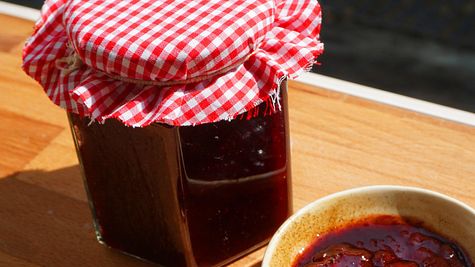 Selbstgemachte Kirschmarmelade im Glas mit rot-weiß kariertem Stoffdeckel, daneben eine Schale mit frischer Marmelade auf einem Holzbrett in der Sonne - Foto: Anne Kurz / LECKER.de