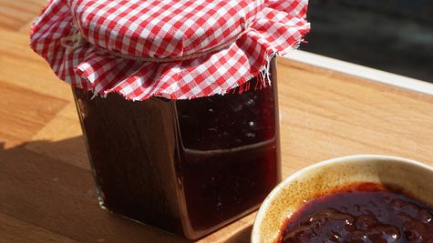Selbstgemachte Kirschmarmelade im Glas mit rot-weiß kariertem Stoffdeckel, daneben eine Schale mit frischer Marmelade auf einem Holzbrett in der Sonne - Foto: Anne Kurz / LECKER.de