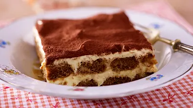 Ein tiefer Teller mit einem Stück Tiramisu steht auf einem Holztisch mit rot-weiß-kariertem Platz-Set  - Foto: LECKER @ Bauer Media Group