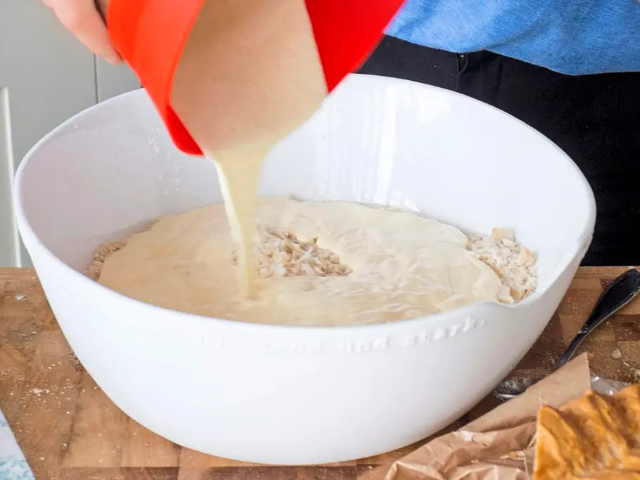 Eine cremige Ricotta-Ei-Mischung wird aus einer roten Schüssel in eine große weiße Schüssel mit Brot gegeben, auf einem Holzbrett liegt daneben ein Stück frisches Weißbrot.