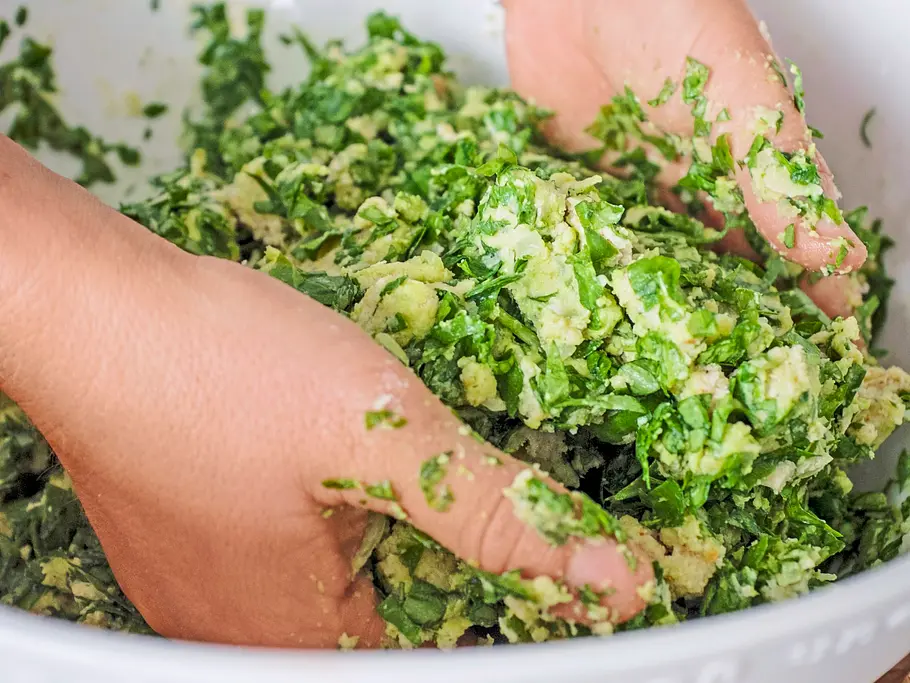 Mit den Händen wird eine grüne Spinat-Brot-Masse in einer großen weißen Schüssel vermengt, bereit zum Formen von Klößen.