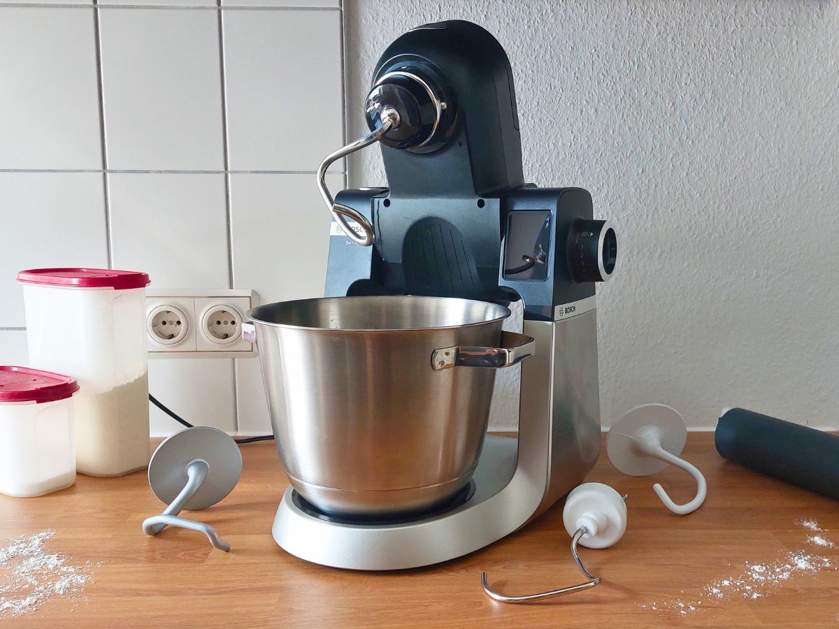 Eine Küchenmaschine mit Knethaken, umgeben von drei anderen Knethaken, Mehl und Zucker sowie einer Teigrolle