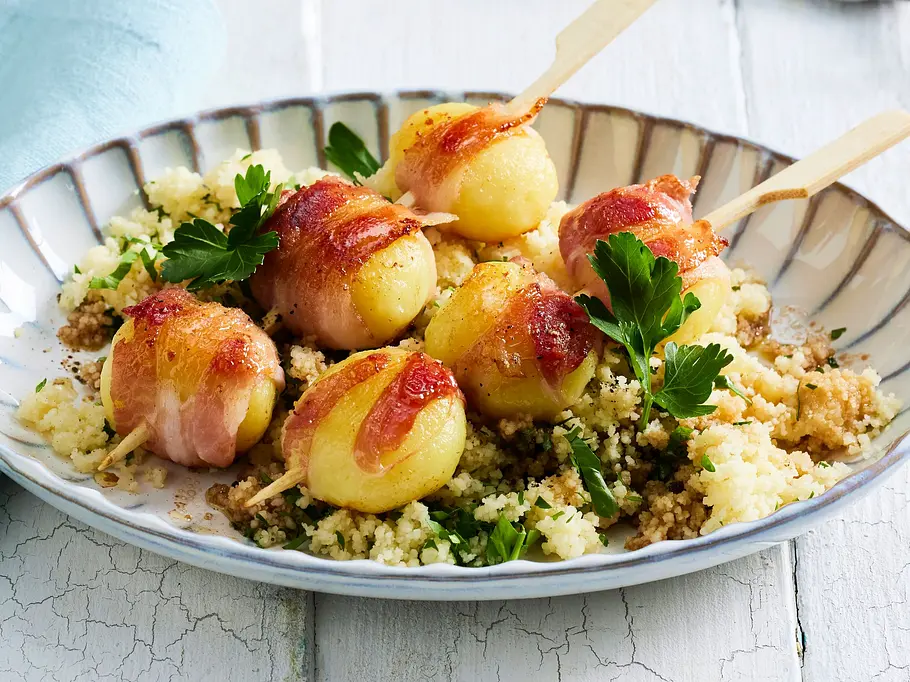 Knödel-Speck-Spieße auf einem Bett aus lockerem Couscous mit frischer Petersilie, angerichtet auf einem geriffelten Teller mit hellblauer Serviette.