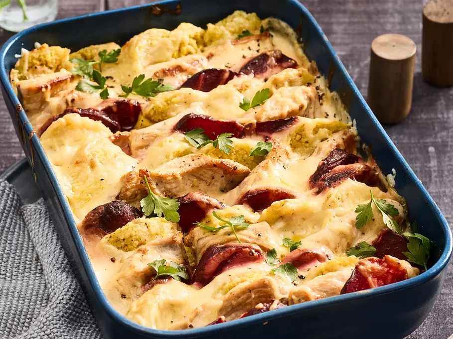 Überbackenes Knödelgratin mit Roter Bete in einer blauen Auflaufform, goldbraun gebacken und mit frischer Petersilie bestreut, serviert auf einem Holztisch mit Küchenutensilien.