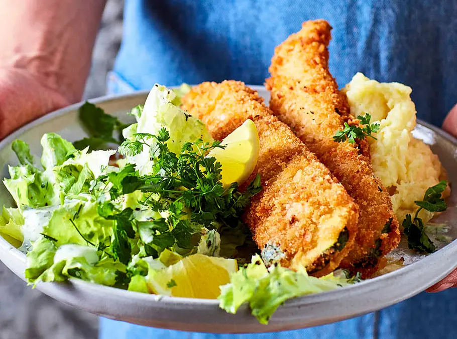 Zwei goldbraun panierte Zucchini-Schnitzel auf einem Teller mit grünem Salat, Zitronenspalten und cremigem Kartoffelpüree.