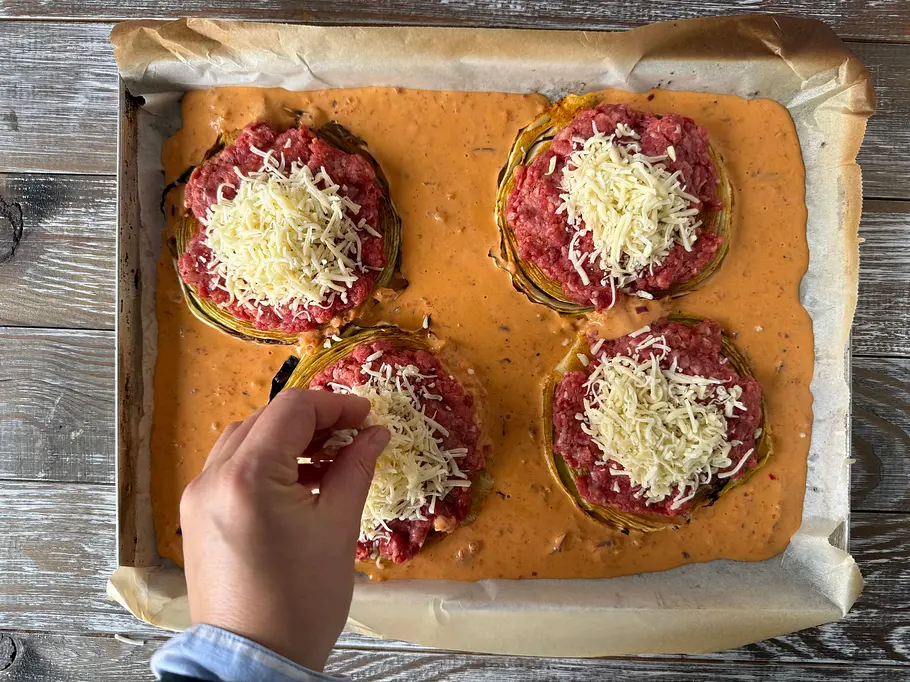 Eine Hand streut Käse auf vier mit Hackfleisch belegte Kohlscheiben, die auf einem Backblech in orangefarbener Soße liegen.