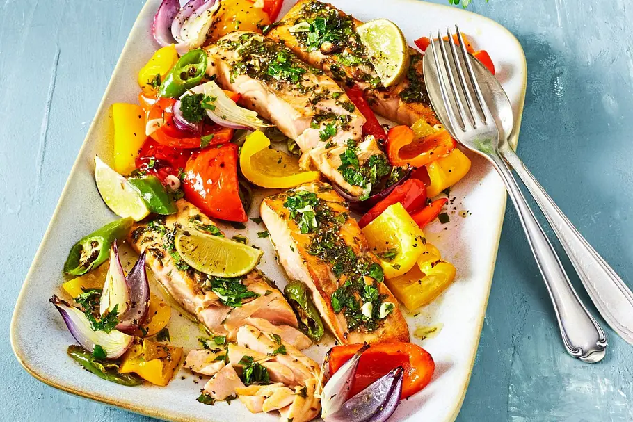 Kräuter-Lachs auf Paprika aus dem Airfryer Kräuter-Lachsfilet in Portionen auf Paprika aus dem Airfryer gebettet auf Paprikagemüse mit gelben und roten Paprikas und roten Zwiebeln. Das Gericht befindet sich auf einer weißen Servierplatte, dekoriert mit Limettenspalten.