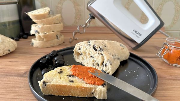 Angeschnittenes Oliven-Ciabatta, von dem eine Scheibe mit rotem Pesto und Olivenöl bestrichen ist, liegt auf einem schwarzen Teller vor dem Krups 3Mix 9000 Handrührgerät - Foto: LECKER.de/Nicole Molitor