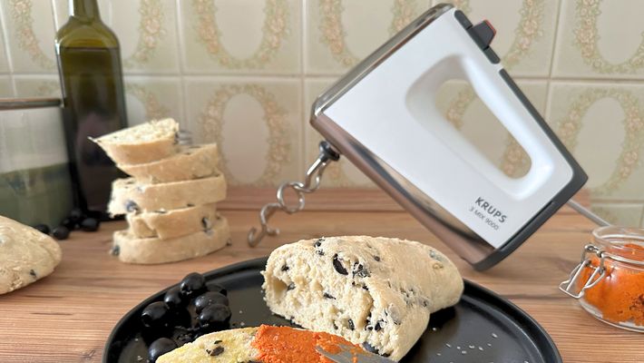 Angeschnittenes Oliven-Ciabatta, von dem eine Scheibe mit rotem Pesto und Olivenöl bestrichen ist, liegt auf einem schwarzen Teller vor dem Krups 3Mix 9000 Handrührgerät - Foto: LECKER.de/Nicole Molitor