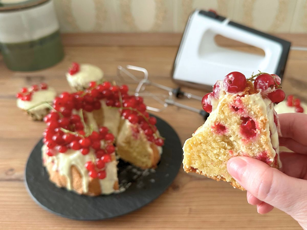 Roter Johannisbeer-Gugelhupf mit einem nah in die Kamera gehaltenen Stück Kuchen und dem Krups 3Mix 9000 etwas unscharf im Hintergrund