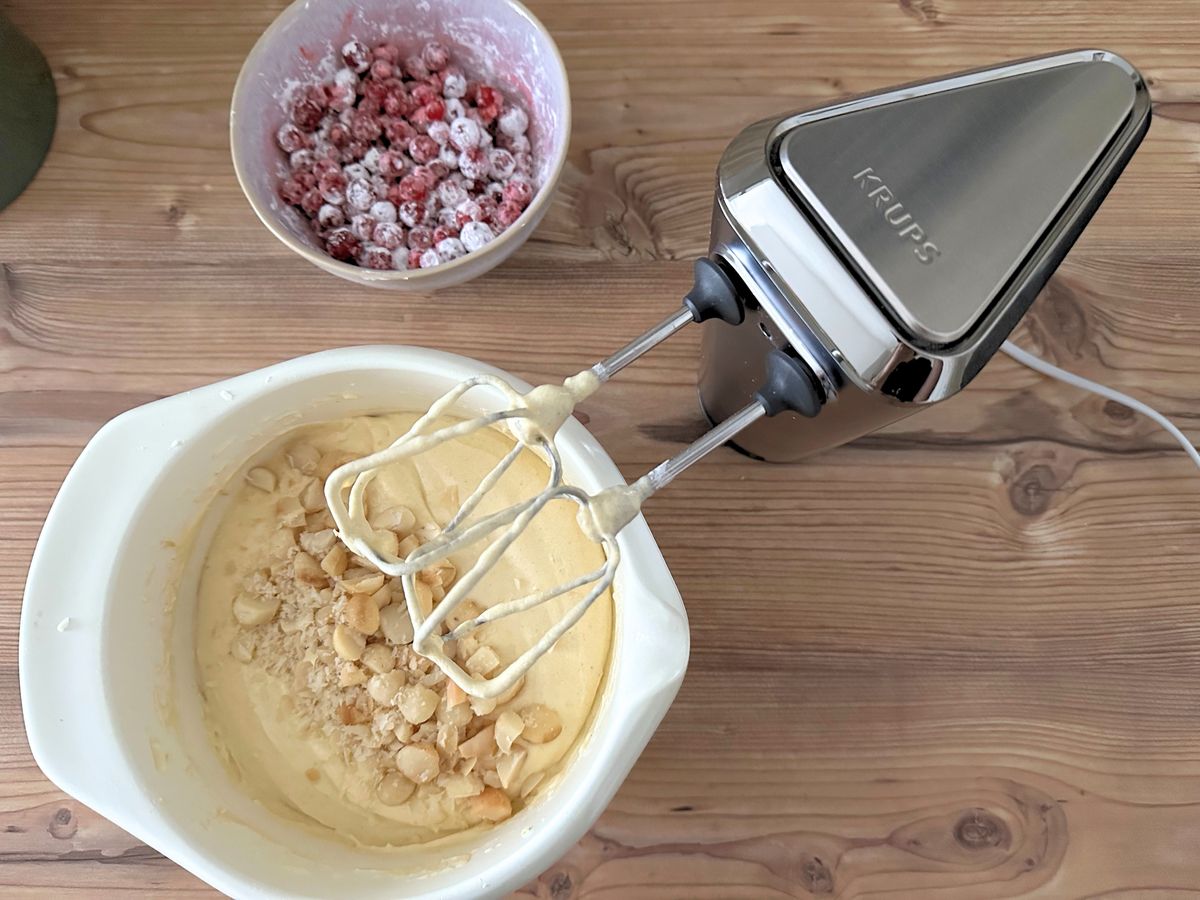 Topshot von einer Schüssel mit Rührteig und grob gehalten Macadamias, daneben der Krups-handmixer samt Quirlen und eine Schale mit bemehlten roten Johannisbeeren