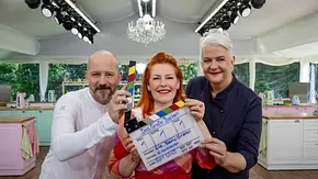 Drei Personen stehen im Backzelt von „Das große Backen“ vor pastellfarbenen Küchenzeilen und halten gemeinsam eine Filmklappe mit der Aufschrift „Das große Backen“ in die Kamera; im Hintergrund sind Kronleuchter und Backstationen zu sehen. - Foto:  Joyn / Claudius Pflug