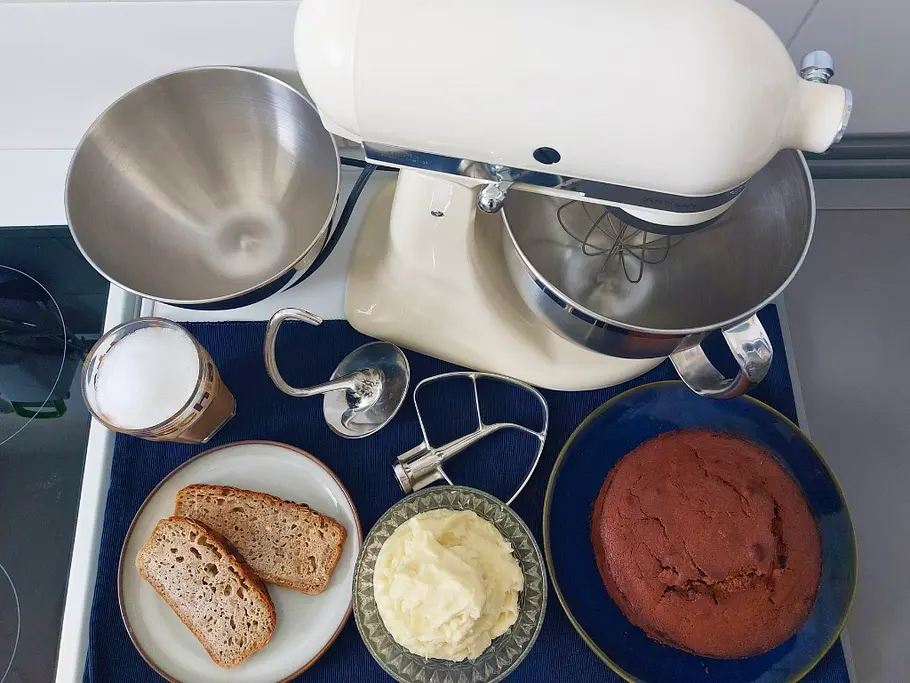Küchenmaschine Vergleichstest Man sieht eine cremefarbene KitchenAid-Küchenmaschine mit Rührschüssel und Teigschabern von oben auf einer Küchenanrichte neben separaten Tellern mit frisch gebackenen Brotscheiben, Kartoffelpüree und Schokokuchen.