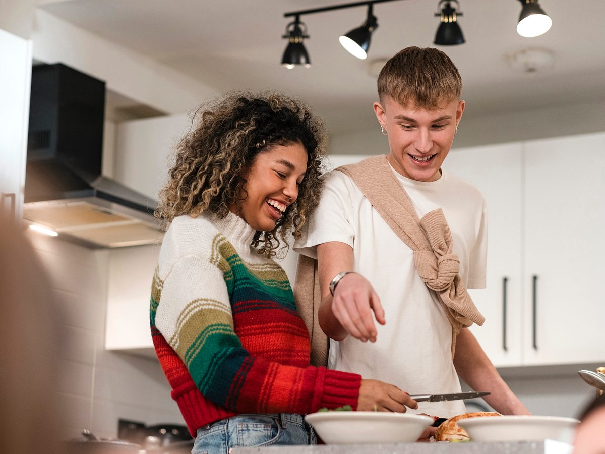 Zwei junge Menschen stehen nebeneinander in einer Küche und bereiten lächelnd eine Mahlzeit an der Arbeitsplatte zu.