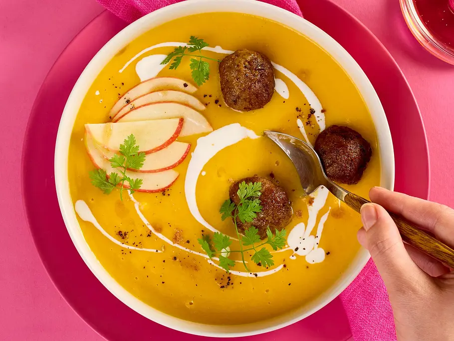 Cremige Kürbis-Apfel-Suppe in einer weißen Schale, garniert mit drei Fleischbällchen, frischen Apfelscheiben, Kräutern und einem Klecks Sahne, serviert auf pinkem Teller mit passender Serviette und Glas.