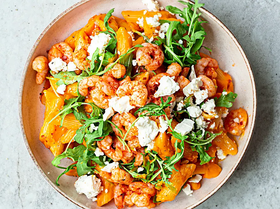Kürbis-Pasta mit gebratenen Garnelen, zerbröseltem Feta und frischem Rucola in einer hellen, runden Schale.