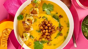 Cremige Kürbis-Süßkartoffel-Suppe in einer weißen Schale, garniert mit knusprigen Kichererbsen, frischem Koriander und einem gegrillten Hähnchenspieß, serviert mit pinker Serviette und farbigem Teller. - Foto: LECKER @ Bauer Media Group