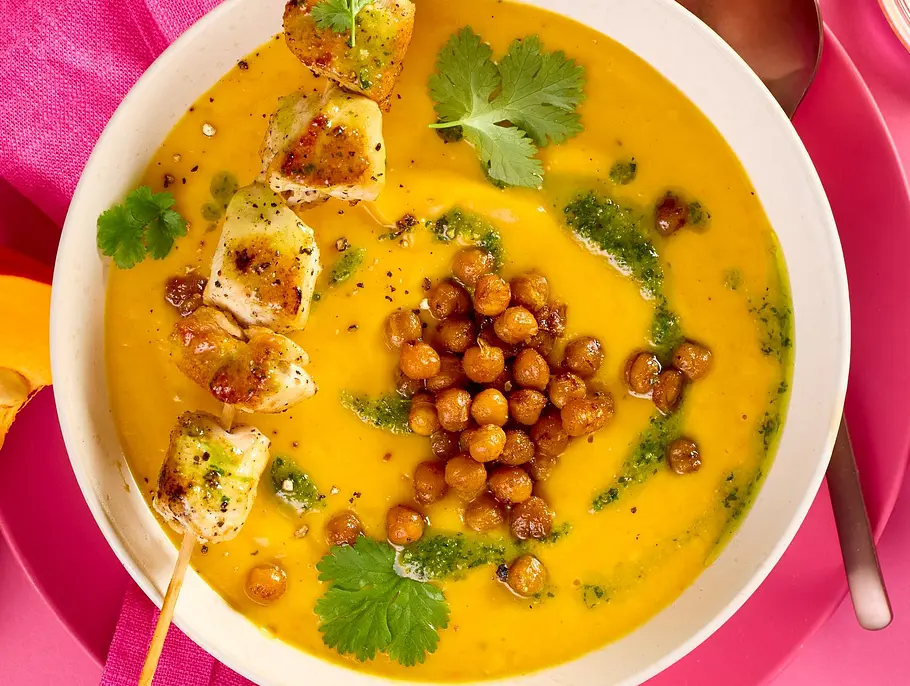 Cremige Kürbis-Süßkartoffel-Suppe in einer weißen Schale, garniert mit knusprigen Kichererbsen, frischem Koriander und einem gegrillten Hähnchenspieß, serviert mit pinker Serviette und farbigem Teller.