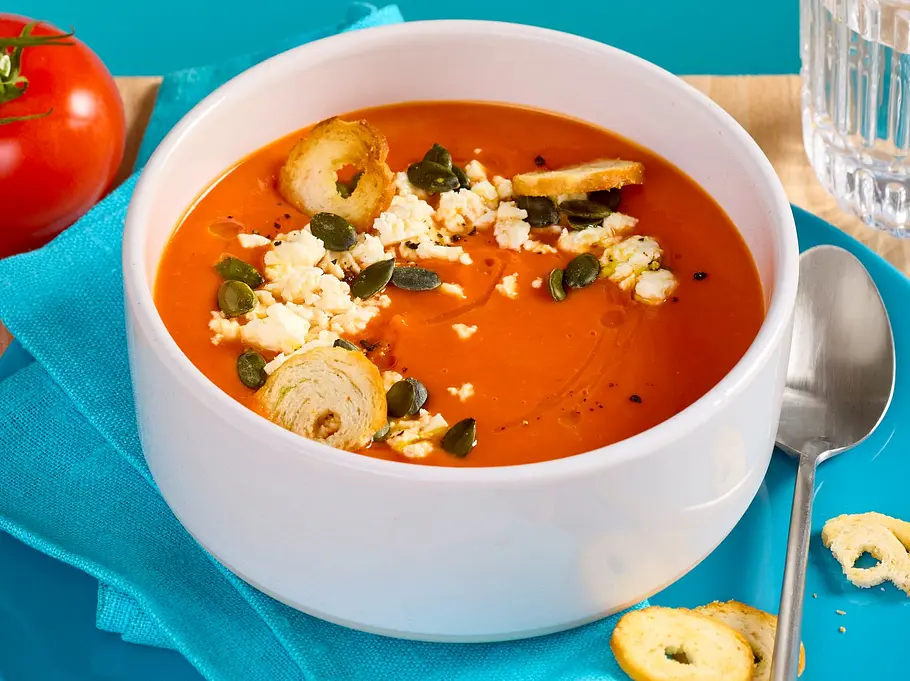 Kürbis-Tomaten-Suppe in einer weißen Schale, garniert mit zerbröseltem Feta und Kernen, serviert auf einem blauen Teller mit Serviette, Croutons und einem Glas Wasser.