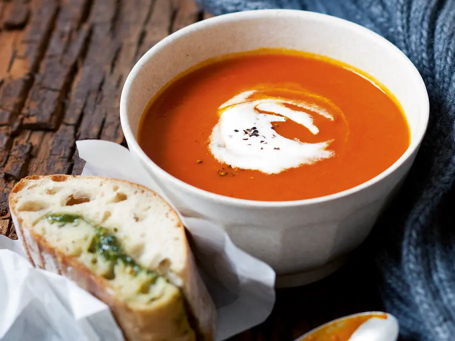 Eine Schale Kürbis-Tomatensuppe mit Sahnetopping und Brotstück auf einem Holztisch serviert.