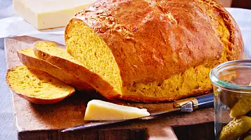 Ein rundes, goldgelbes Kürbisbrot liegt angeschnitten auf einem Holzbrett, daneben Käse und ein Glas mit eingelegten Gurken. - Foto: LECKER @ Bauer Media Group