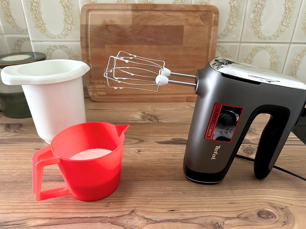 Der Tefal Handmixer steht auf einer hölzernen Arbeitsfläche neben einem großen und einem Kleinen Rührbecher mit Sahnetropfen an den Quirlen