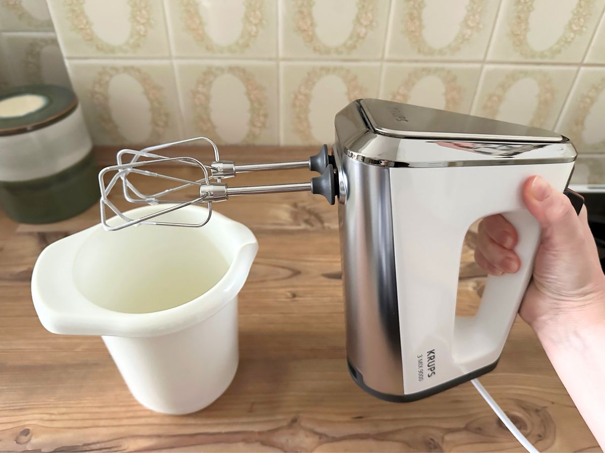 Kuchenteig mit Macadamias im Rührtest vom rups-Handmixer Der Krups 3Mix 9000 Handmixer wird von einer Hand gehalten, sodass sichtbar ist, dass die Quirle nur halb mit Sahne aus dem Becher daneben behaftet sind.
