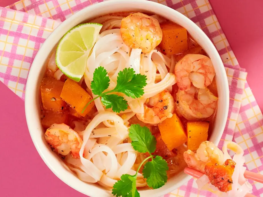 Laksa-Nudelsuppe in weißen Schalen mit Reisnudeln, Garnelen, Butternusskürbiswürfeln, frischem Koriander und Limetten, serviert auf buntem Untergrund mit Essstäbchen.
