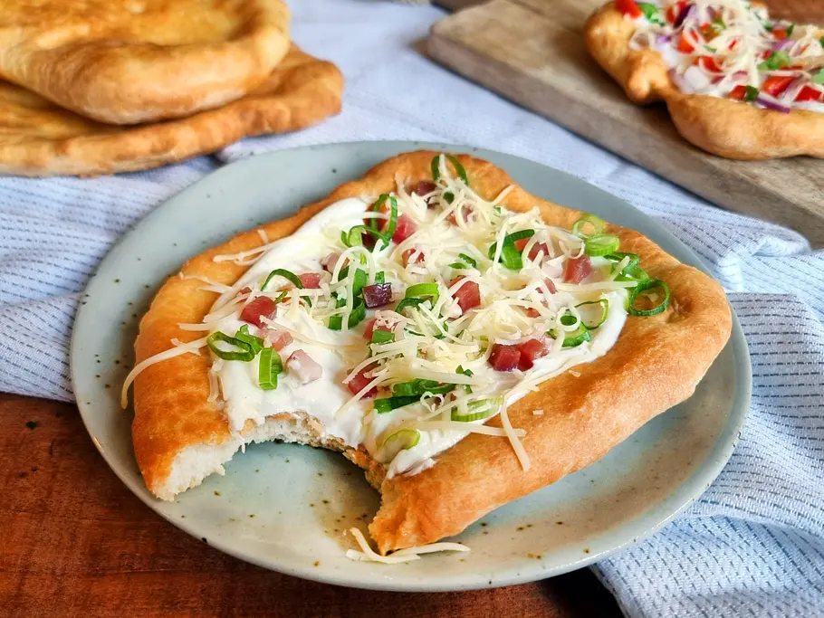 Ungarische Langos: Teigfladen mit weißer Creme, Käse, Lauchzwiebel und Speck belegt.