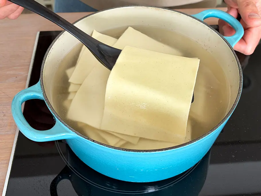 Lasagneblätter kochen In einem blauen Kochtopf werden mehrere Lasagneplatten gekocht. Eine Platte wird mit einem Kochlöffel herausgehoben.