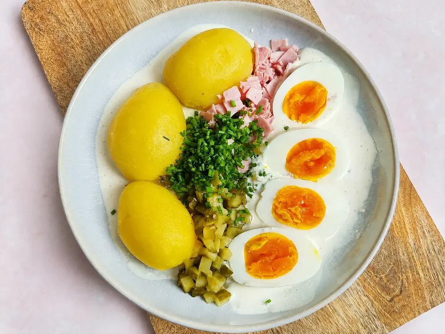 Lazy Kartoffelsalat klassisch Rezept Alle Zutaten des Lazy Kartoffelsalats auf einem Teller: Joghurt-Dressing, Kartoffeln, Gewürzgurken, Kochschinken und gekochte Eier.