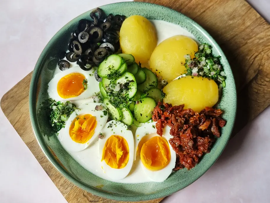 Mediterraner Lazy Kartoffelsalat Die Zutaten für Lazy Kartoffelsalat auf Dressing auf einem Teller: Kartoffeln, Gurken, Kräuter, Oliven, Eier und getrocknete Tomate.