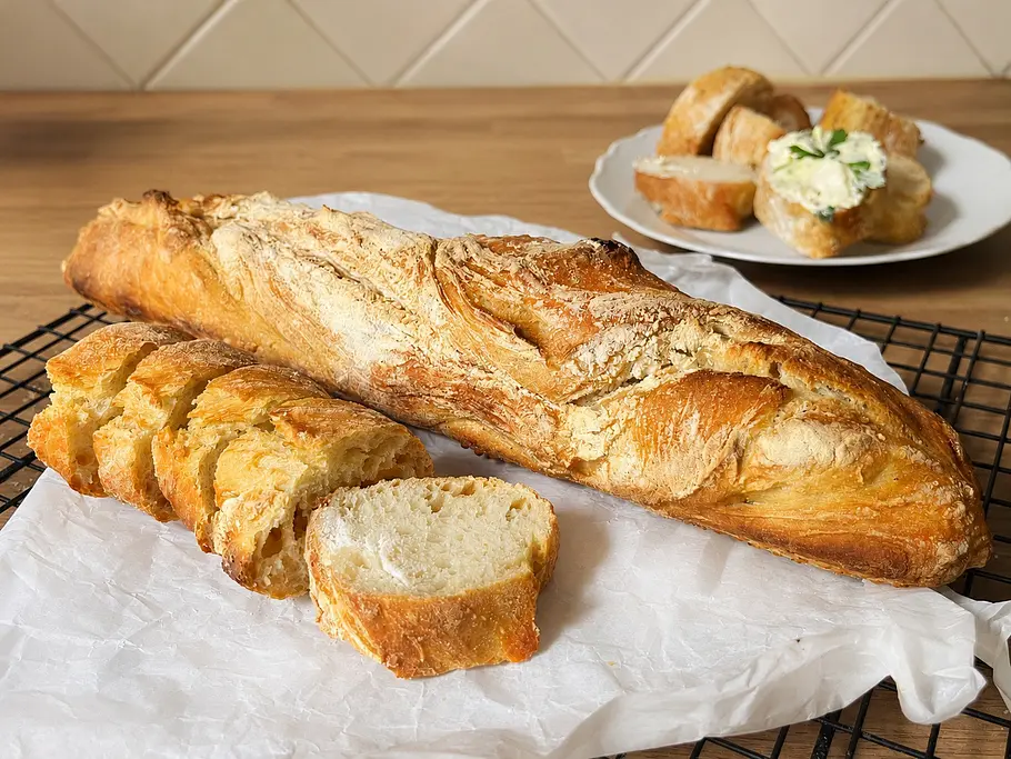Einfaches Baguette ohne Kneten Knuspriges Baguette ohne Kneten auf einem Baguetteblech gebacken, angeschnitten auf Backpapier mit Kräuterbutter im Hintergrund.