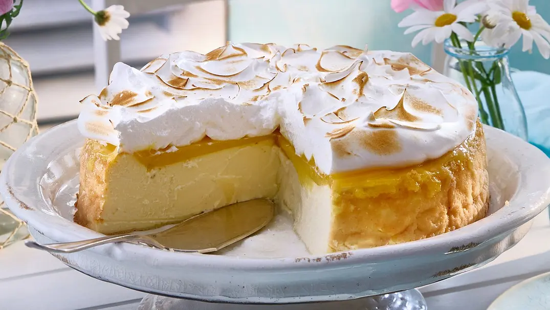 Lemon-Curd-Käsekuchen mit goldbrauner Baiser-Haube auf einer Tortenplatte, daneben einzelne Stücke auf hellblauen Tellern auf einem weiß gedeckten Tisch. - Foto: LECKER @ Bauer Media Group