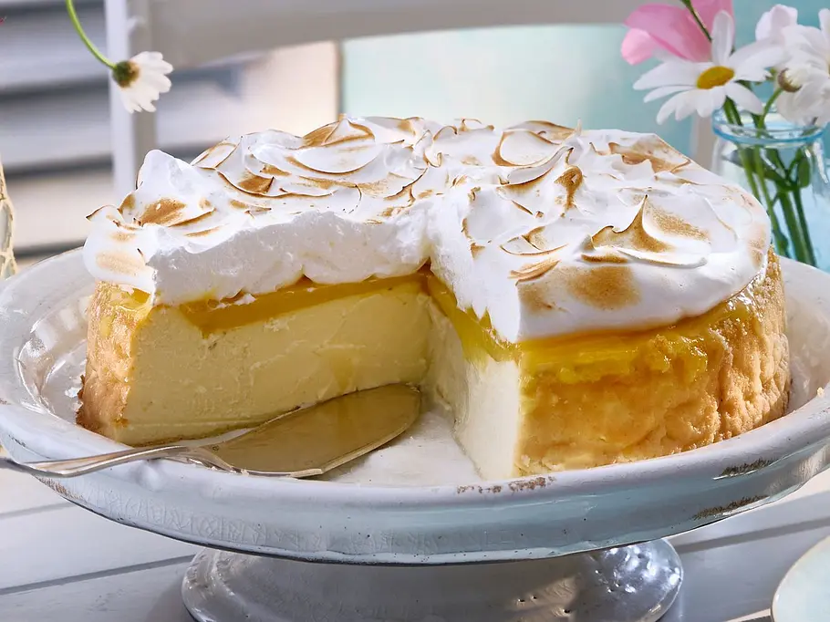 Lemon-Curd-Käsekuchen mit goldbrauner Baiser-Haube auf einer Tortenplatte, daneben einzelne Stücke auf hellblauen Tellern auf einem weiß gedeckten Tisch.